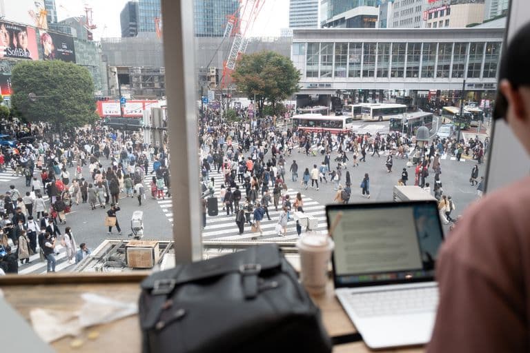 View of a busy city intersection filled with many pedestrians crossing the street, with tall buildings and construction cranes in the background, seen from inside a building with a person working on a laptop at a table.