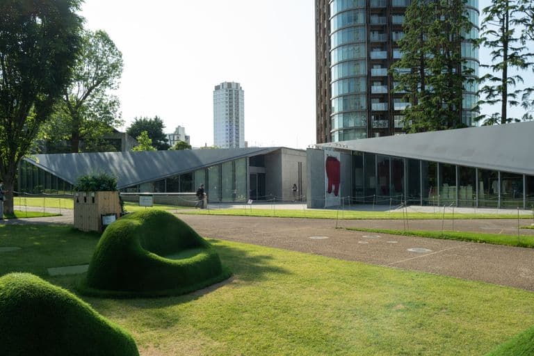 Modern building with large glass windows and sloped roof, surrounded by a park with green grass, trees, and sculpted bushes, in an urban area with tall buildings in the background.