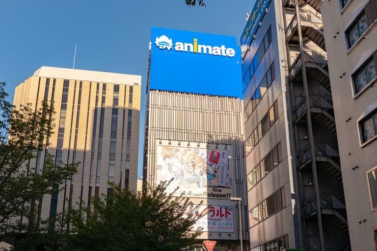 Tall buildings in an urban area with signs, including a large blue billboards reading 'animate' and another with anime character artwork, set against a clear blue sky.
