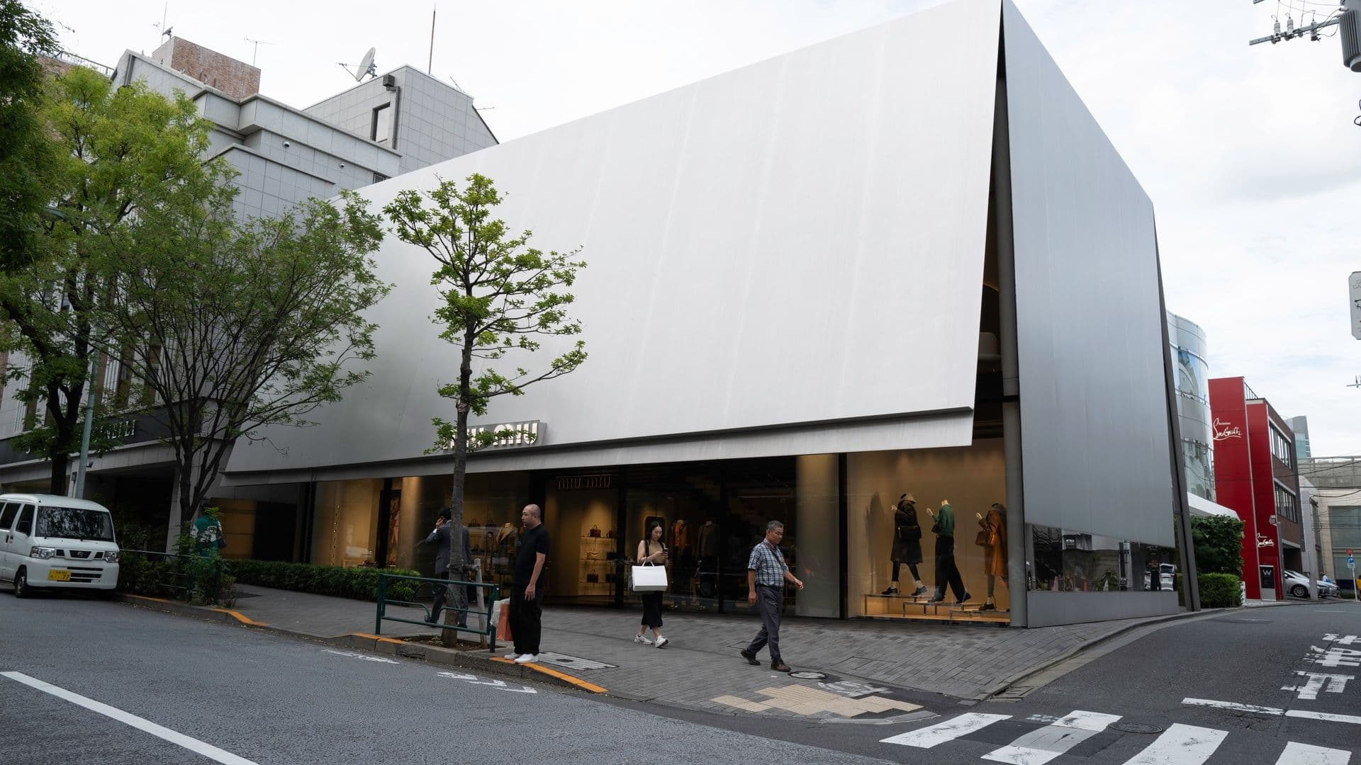 Modern retail store with a sleek, angular white exterior, large glass windows displaying mannequins and clothing, pedestrians walking on the sidewalk, trees, and parked cars in an urban setting.