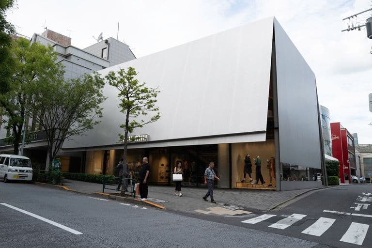 Modern commercial building with a sleek, angled white facade, large glass windows displaying mannequins, and people walking on the sidewalk outside in an urban area.