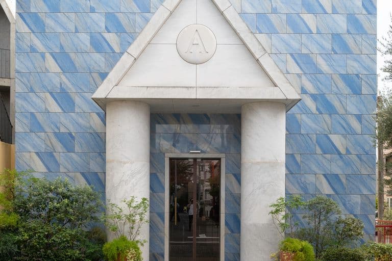 Front view of a modern building with blue tiled walls, red framed windows, and white marble columns supporting a triangular pediment with the letter 'A' engraved, surrounded by potted plants and bushes.