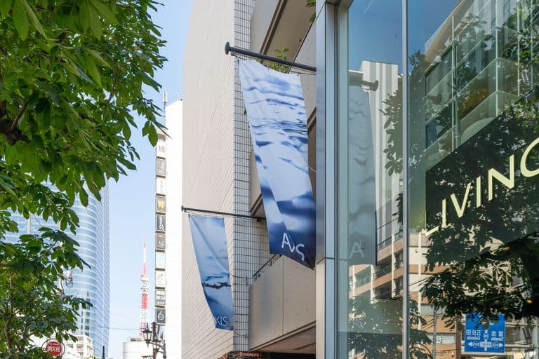 City street with modern buildings, flags with blue and white water design, large glass windows, and a tree canopy.