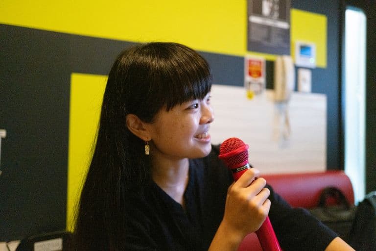 A young woman with long black hair and earrings holding a red microphone, speaking or smiling in a room with colorful background.