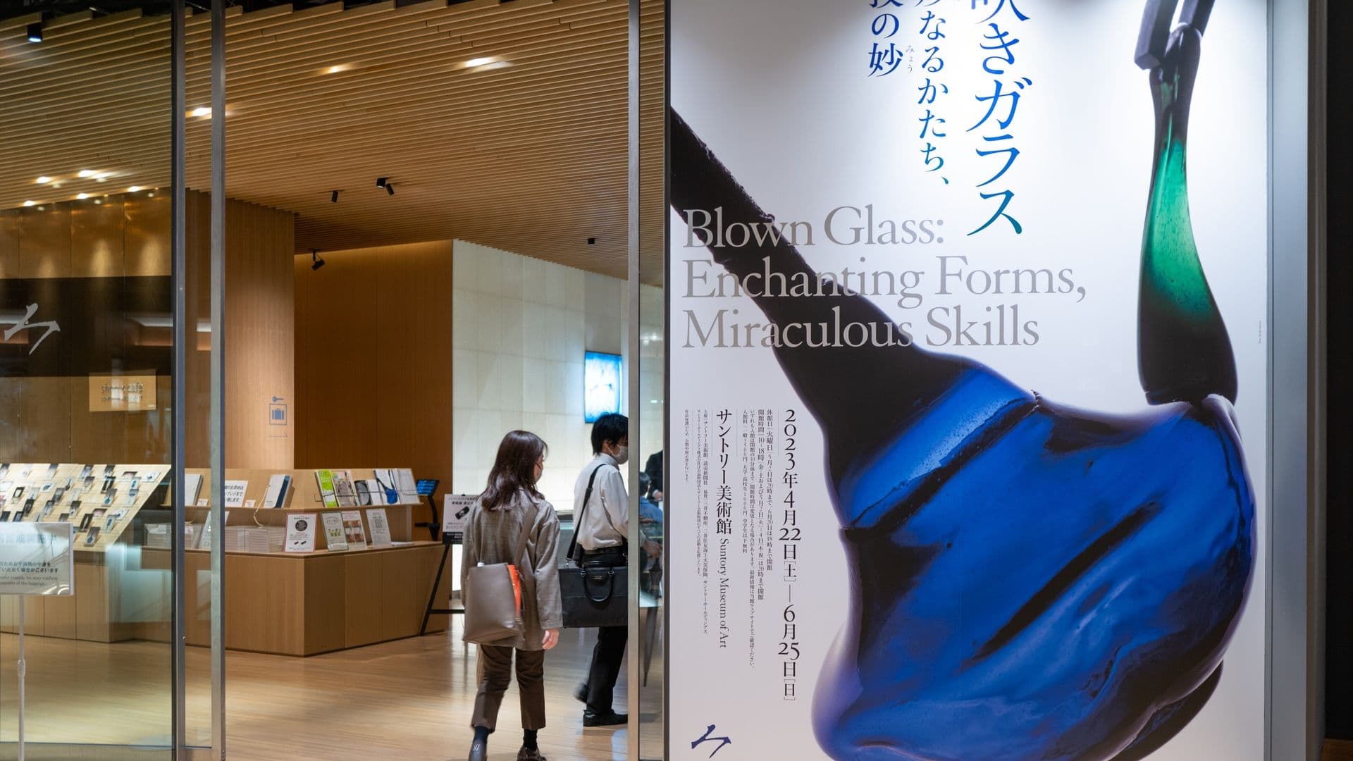 People entering an art museum through glass doors, with a large poster displaying a blown glass sculpture and text about an exhibit titled 'Blown Glass: Enchanting Forms, Miraculous Skills'.