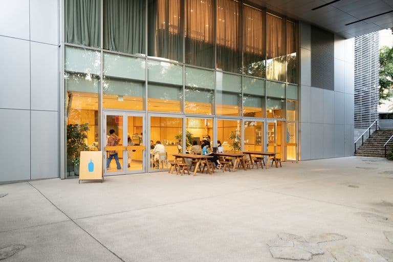 Modern cafe with large glass windows, outdoor seating with wooden benches, and inside there are people sitting and working, illuminated with warm lighting.