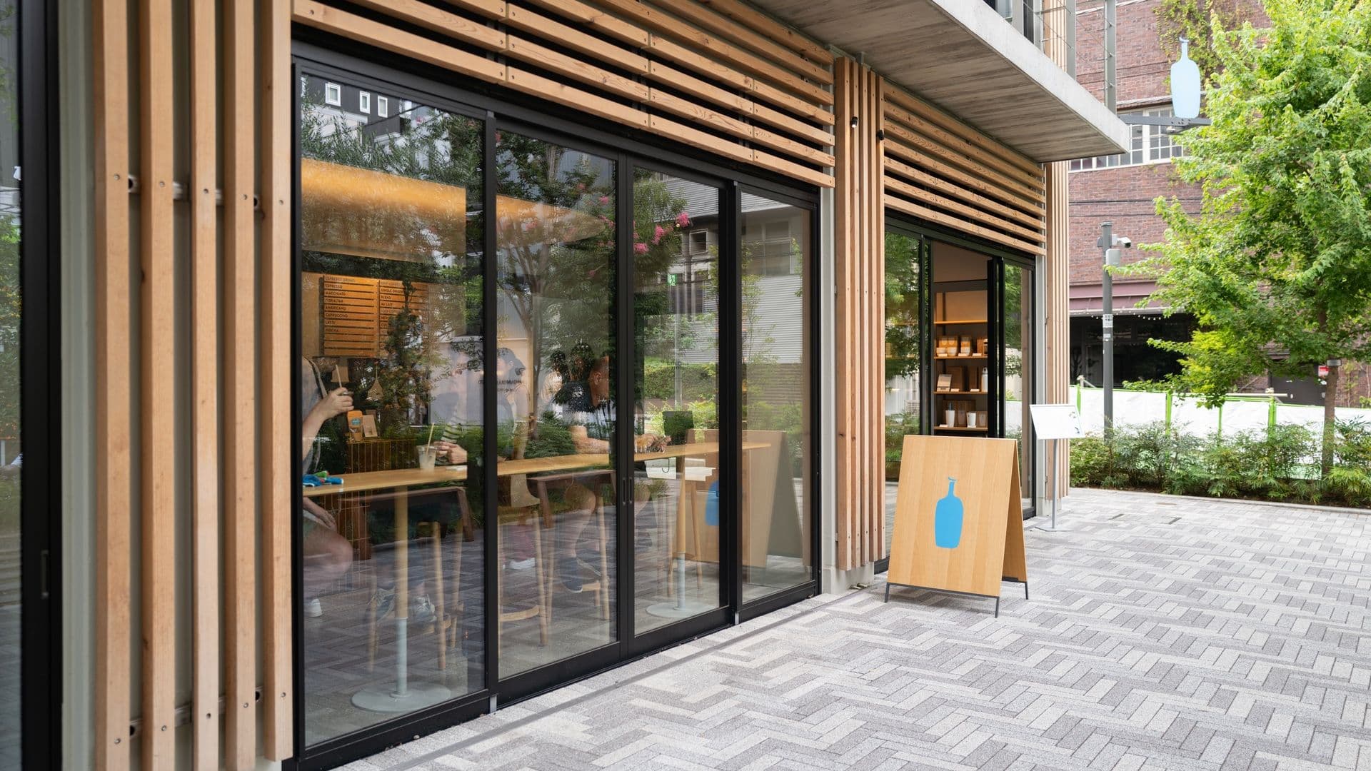 Exterior of a modern building with large glass sliding doors, wooden slats on the facade, and an outdoor paved area with a wooden sign featuring a blue bottle icon, trees, and neighboring buildings in the background.