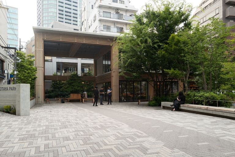 City plaza with modern building and trees, people walking and sitting, glass windows, urban environment.