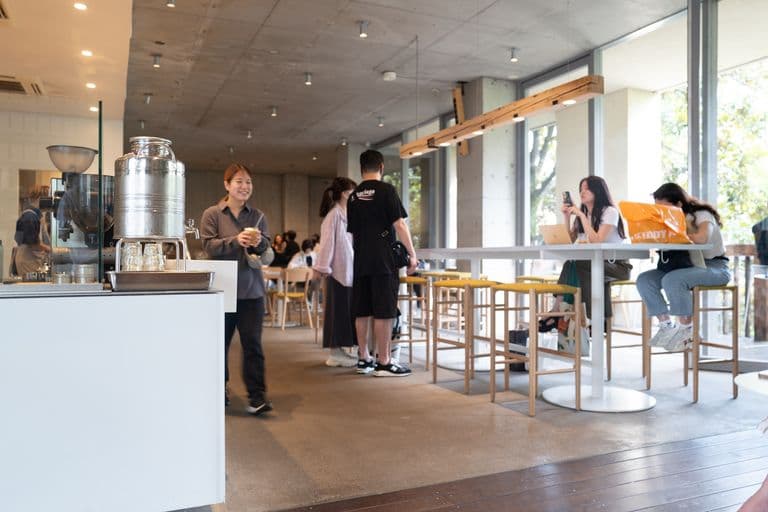 Interior of a modern coffee shop with several customers, some standing in line, some sitting and using their phones, with large windows and natural lighting.
