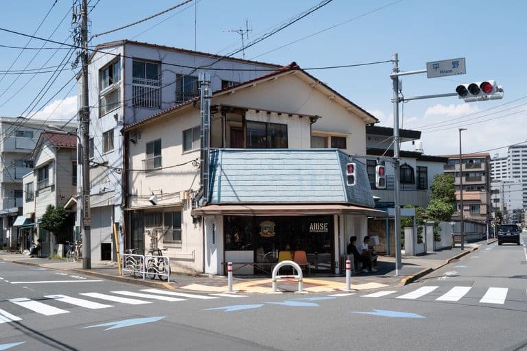 Street corner with a white building housing a coffee shop named 'ARISE', people sitting outside, traffic lights, crosswalks, and electrical wires under a blue sky.