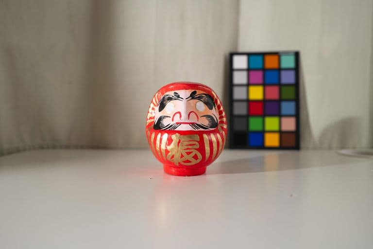 Red Daruma doll with black and gold details on a white surface, with a color calibration chart in the background.