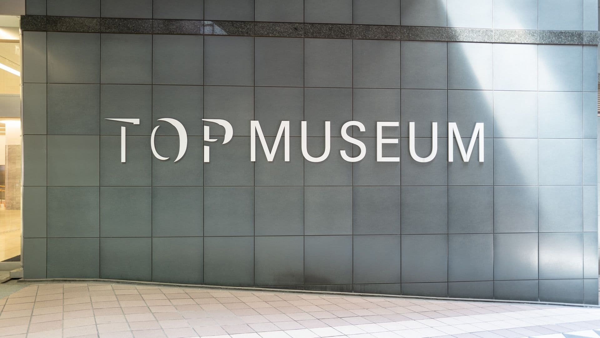 Sign on a wall that reads 'TOP MUSEUM' in large white letters, located outdoors with tiled flooring in front.
