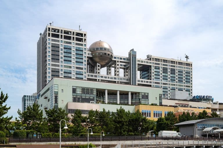 A large modern multi-story building with a prominent metallic sphere structure hanging between two sections of the building. The building has many glass windows and a mix of white, green, and yellow exterior panels. Trees and street lamps are in the foreground, and the sky is partly cloudy.