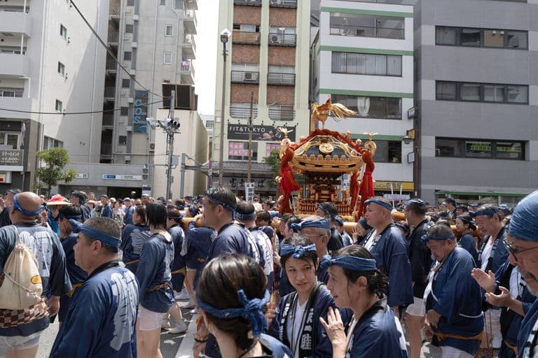 People participating in a traditional Japanese festival parade, carrying a portable shrine (mikoshi), wearing matching blue attire and headbands, in an urban street surrounded by tall buildings.