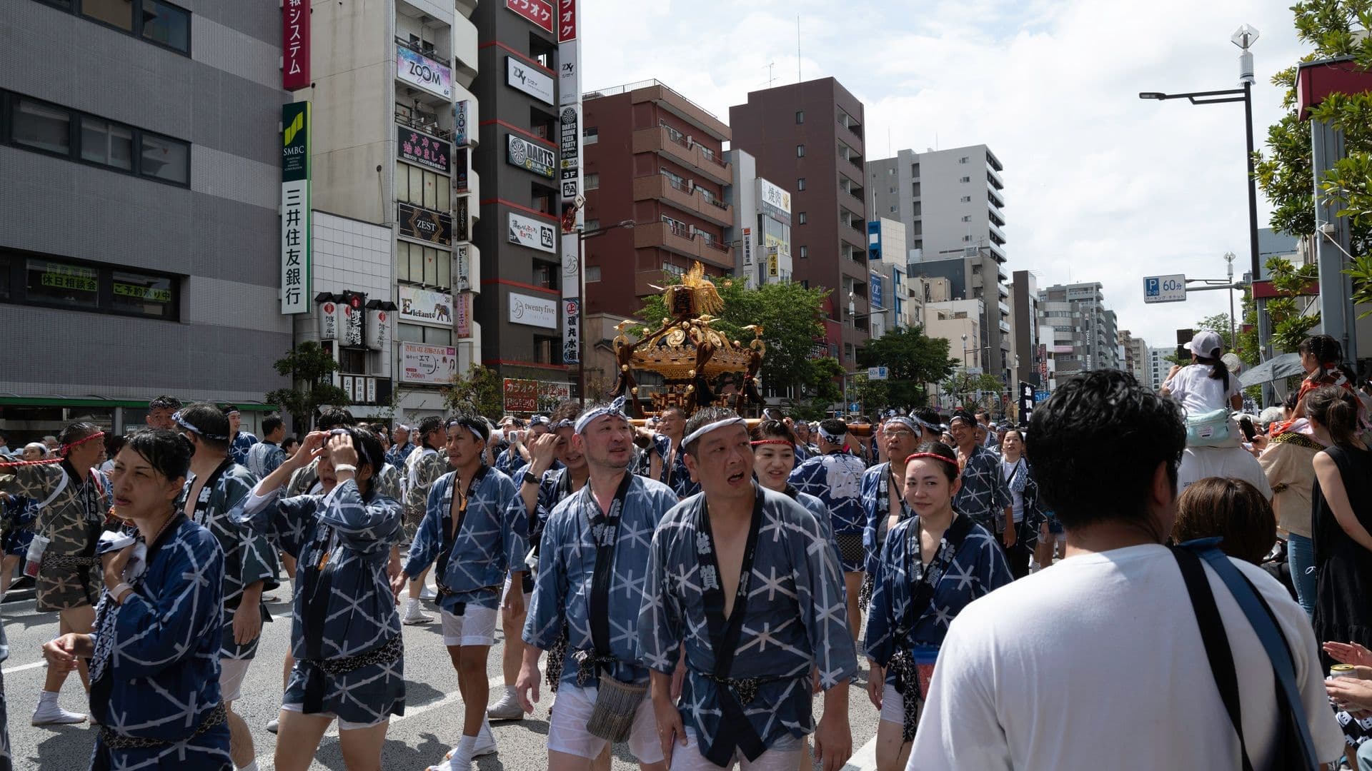 People participating in a traditional festival parade on a city street, carrying a portable shrine (mikoshi) above their shoulders, with tall buildings and signage in the background.