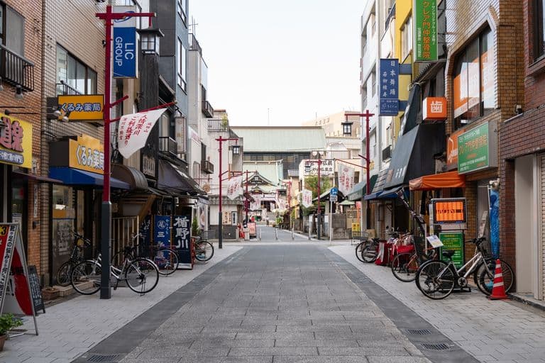An empty pedestrian shopping street with bicycles parked along the sides and various store signs in Japanese, leading towards a traditional building in the background.
