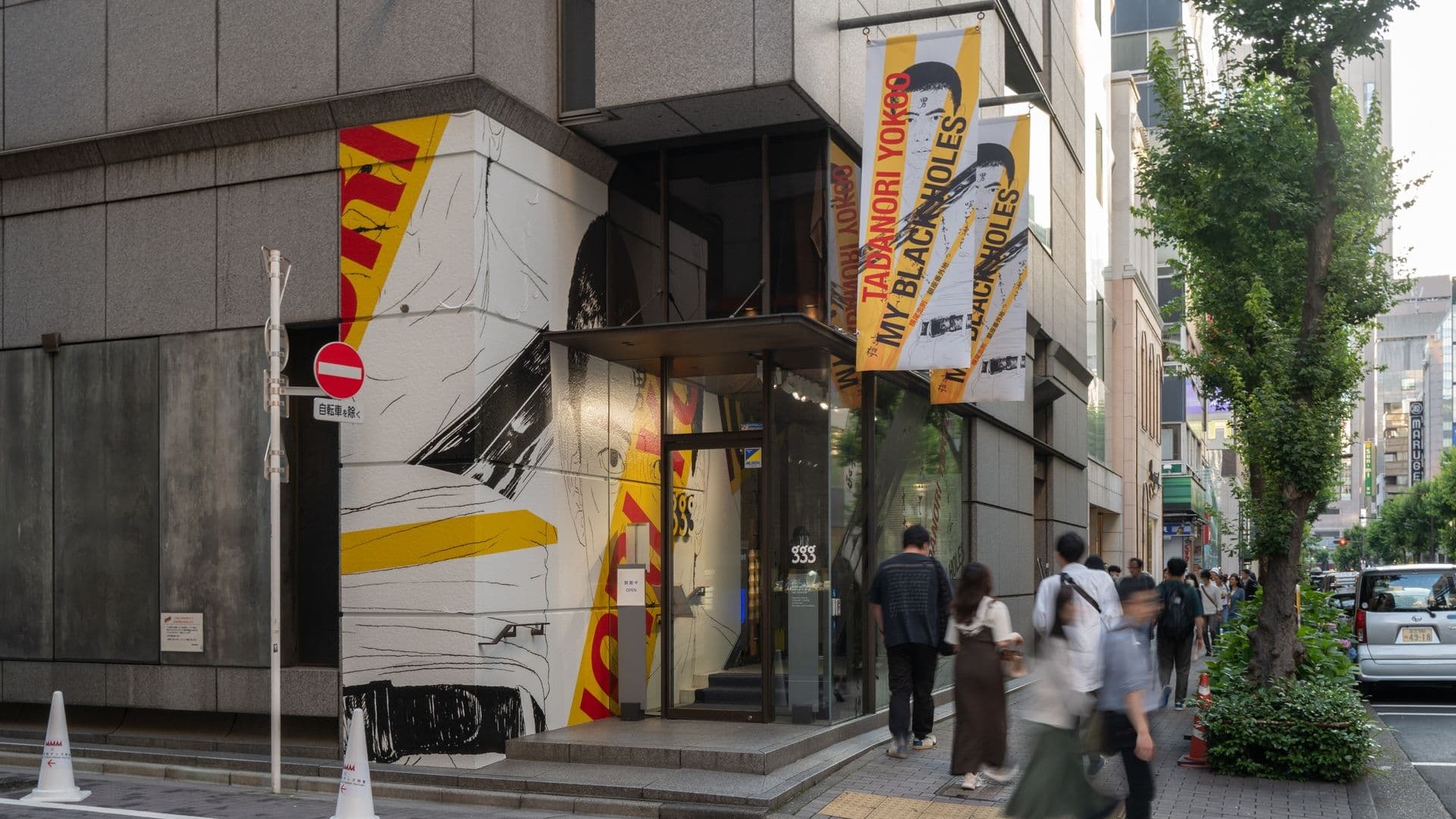 City street scene with a modern building featuring large graphics and banners, pedestrians walking on the sidewalk, and trees lining the street.