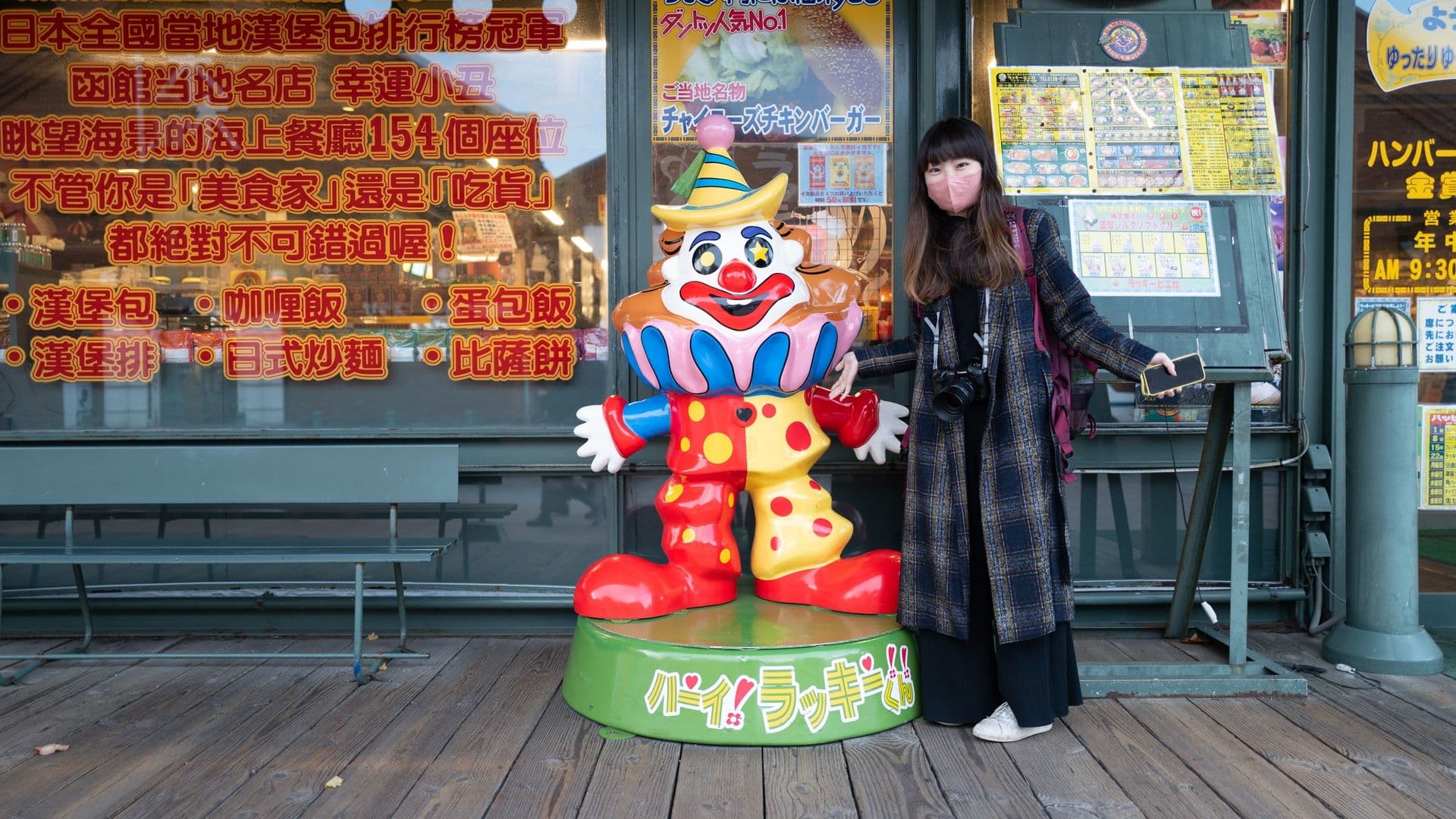 A woman wearing a pink face mask and holding a smartphone stands next to a colorful clown statue in front of a restaurant window with Japanese and Chinese writing. The clown has a party hat, big red shoes, and a vibrant costume with polka dots. The woman has a camera hanging from her neck and a backpack, and she is touching the clown's hand.