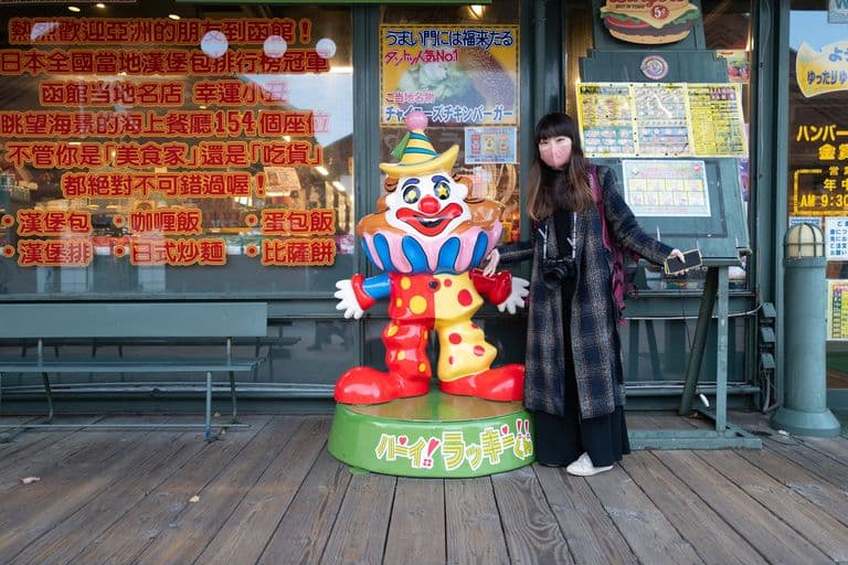 A woman wearing a pink face mask and a plaid coat standing next to a colorful clown statue outside a restaurant with Japanese signage. The woman is holding a camera and a smartphone.
