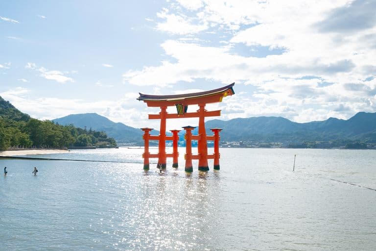 A red Japanese torii gate standing in shallow water with mountains in the background and people swimming nearby.