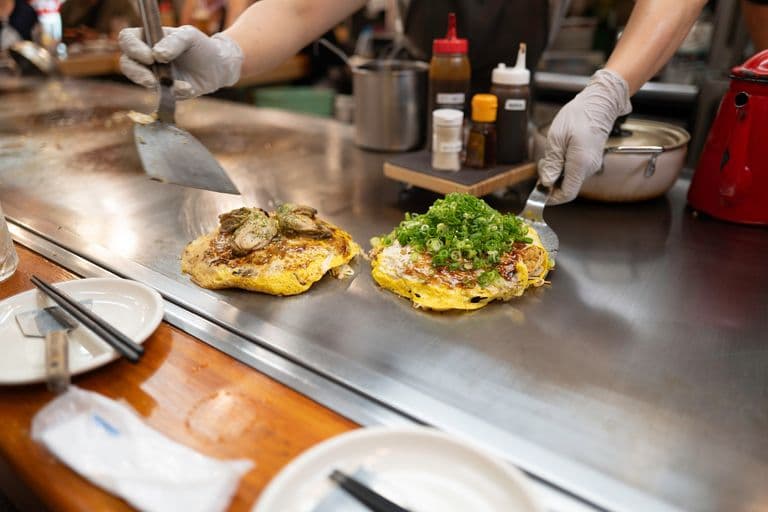 Two chef preparing Japanese-style omelets on a teppanyaki grill, one topped with green onions, and the other with oysters.