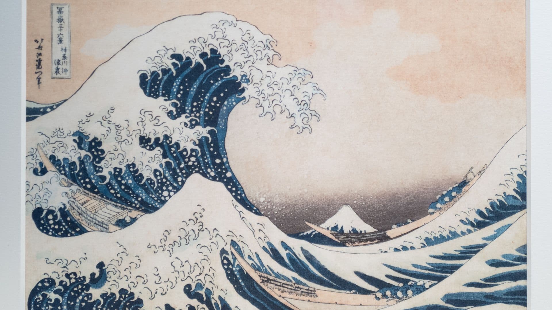 A famous Japanese woodblock print depicting a large, crashing ocean wave with boats near the crest, Mount Fuji in the background, and a pink sky.