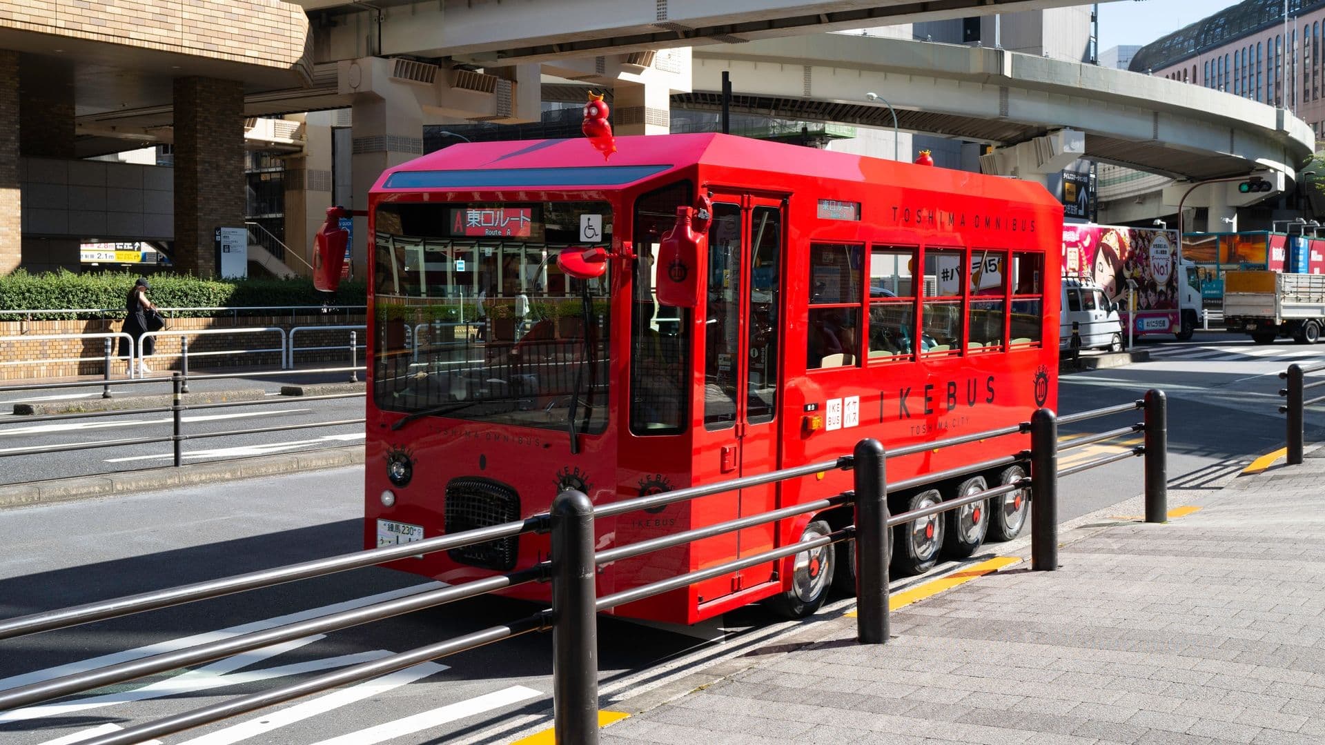 A small red bus designed to look like a train car parked on a city street, with a sign indicating Route A and accessible features, surrounded by traffic barriers and other vehicles in an urban area.