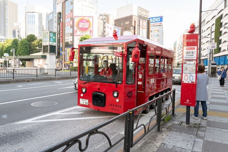 A small red vehicle resembling a bus parked at a bus stop in an urban area with people walking on the sidewalk, tall buildings, and signs in Japanese.