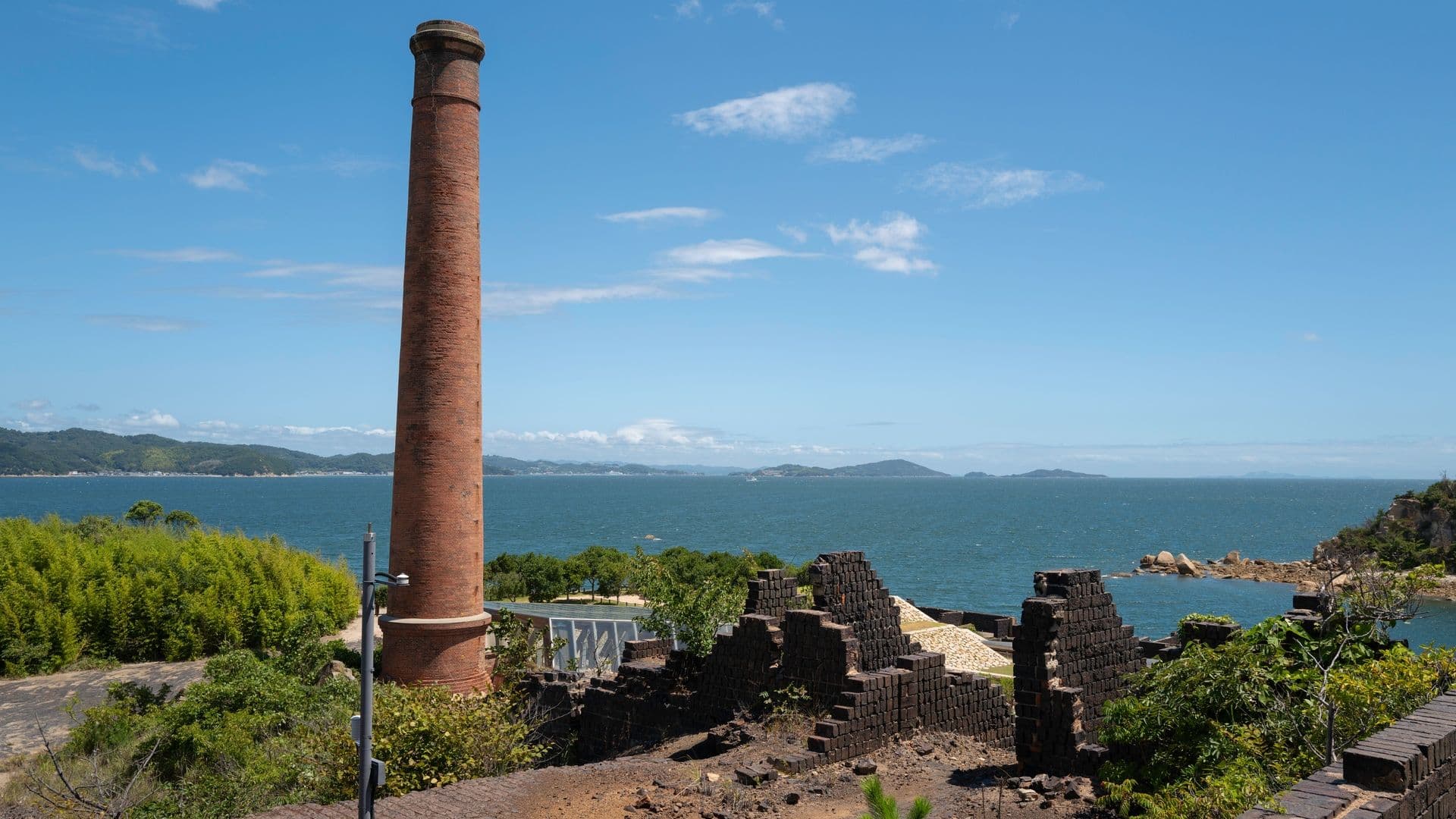 A tall, old brick chimney in front of a body of water with greenery and island in the background, blue sky with some clouds.
