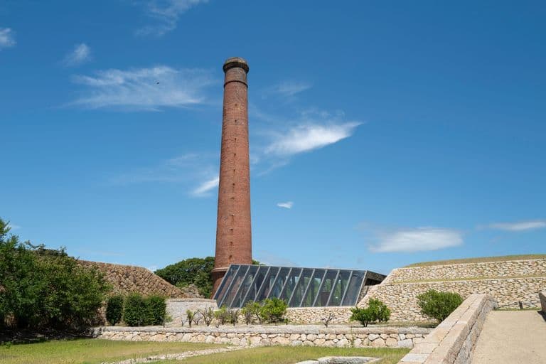 A tall brick smokestack, part of an industrial building, with a modern glass structure in front and green trees and bushes surrounding the area. The sky is blue with some clouds.