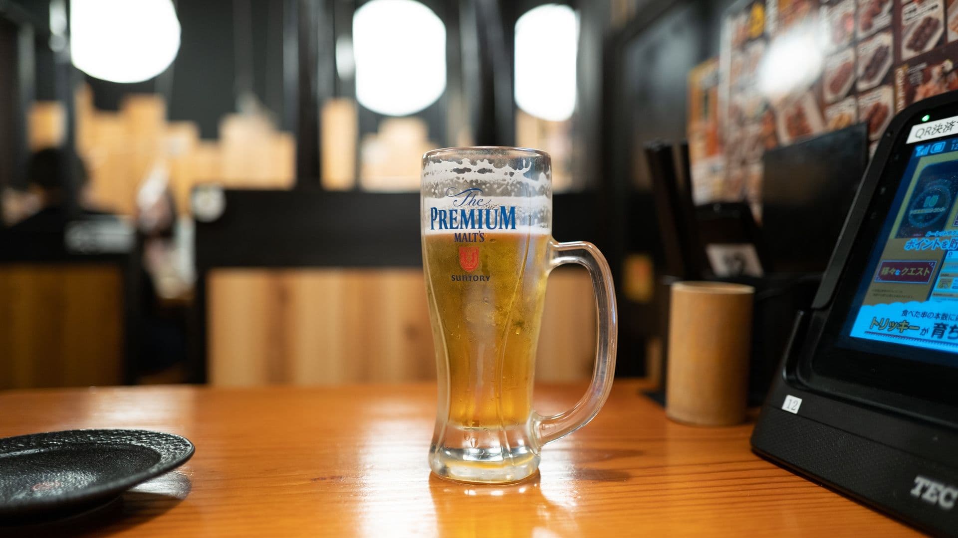 Glass of Suntory Premium Malt's beer on a wooden table in a restaurant with blurred background and digital menu screen.