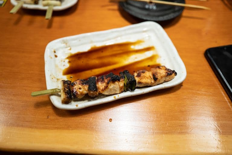 A mostly empty rectangular plate with a grilled chicken skewer and soy sauce on a wooden table in a restaurant setting.