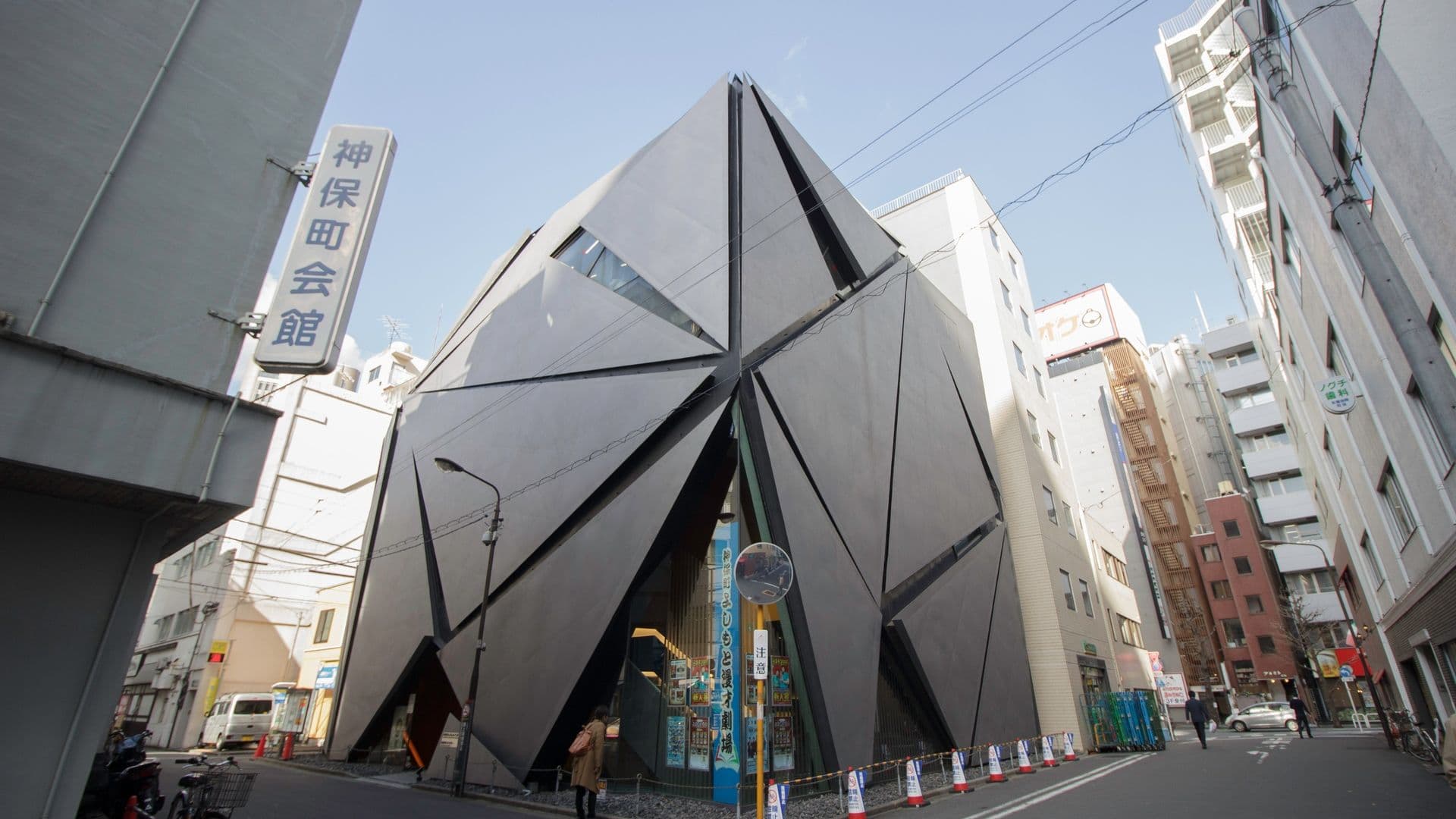 Modern black building with geometric angular facade in a city street surrounded by tall buildings and pedestrians.