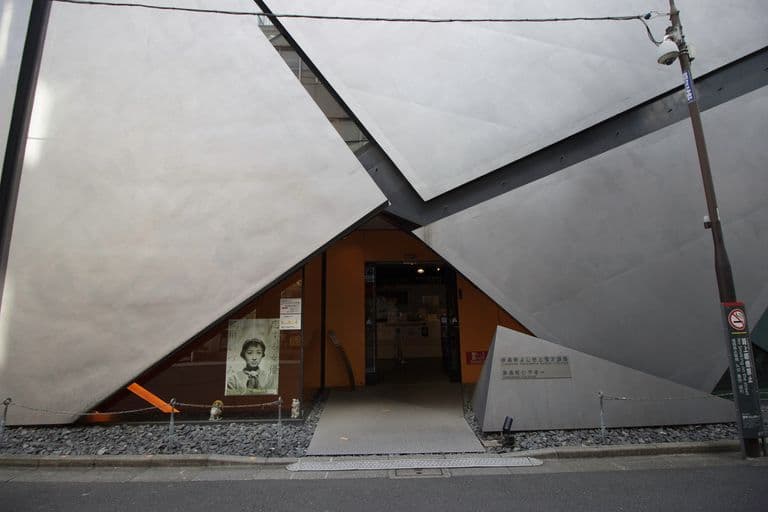 Modern building entrance with angular grey concrete facade, glass door, and a poster with a person's face in a display window.