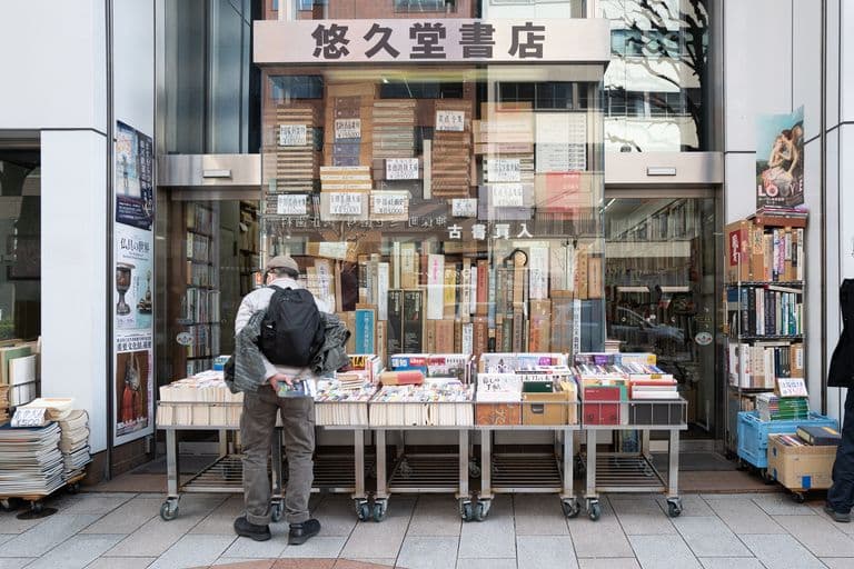 A person shopping for books outside a Japanese bookstore with a display of books on tables and stacked inside the store, with signage in Japanese.