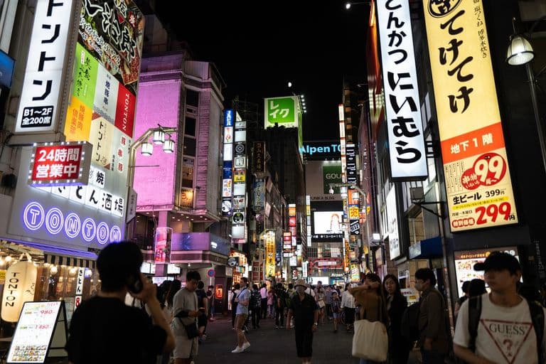 A bustling city street at night filled with bright, colorful neon signs and advertisements in Japanese, with numerous pedestrians walking along the street.
