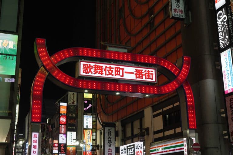 Nighttime view of a busy Japanese street with illuminated signs and a large red archway sign.