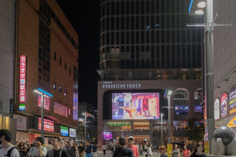 Night view of Kabukicho Tower in Tokyo, with a large digital billboard displaying a colorful image, surrounded by a crowd of people walking outdoors.