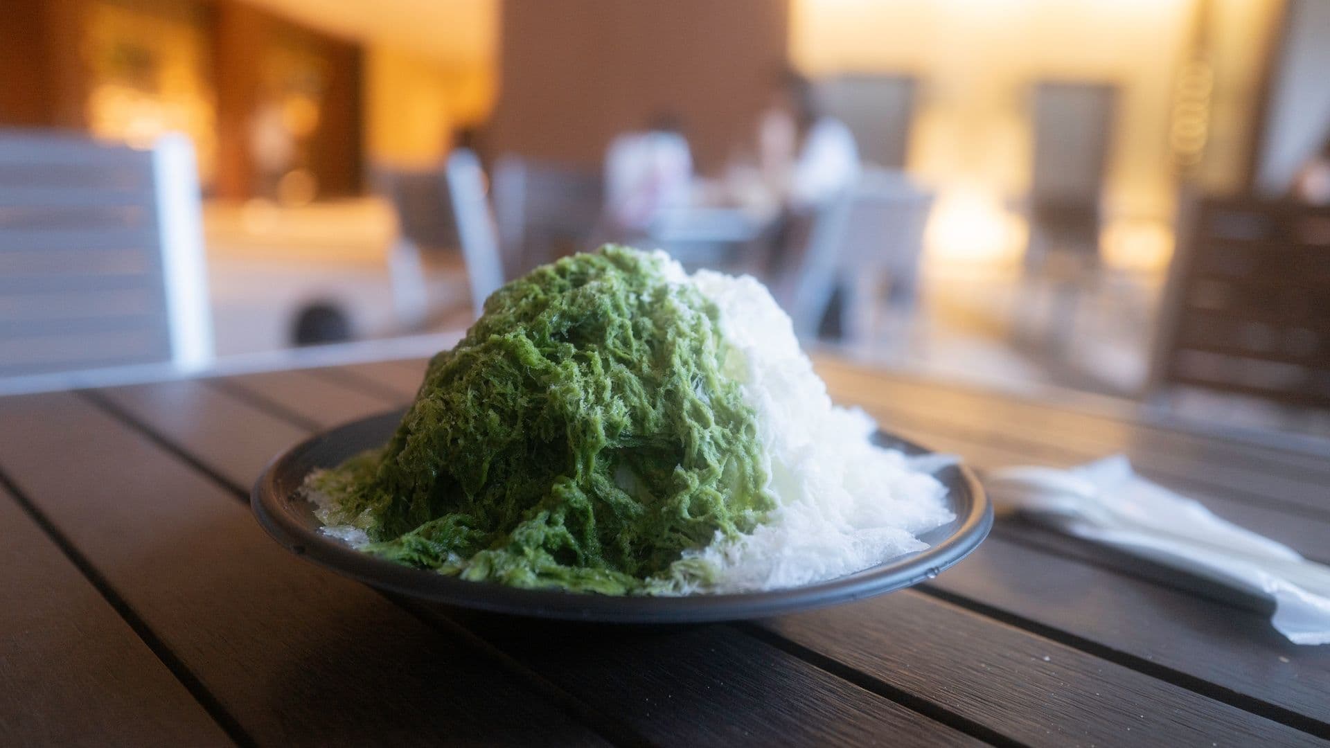 Plate of green and white shaved ice dessert on wooden table in a restaurant setting.