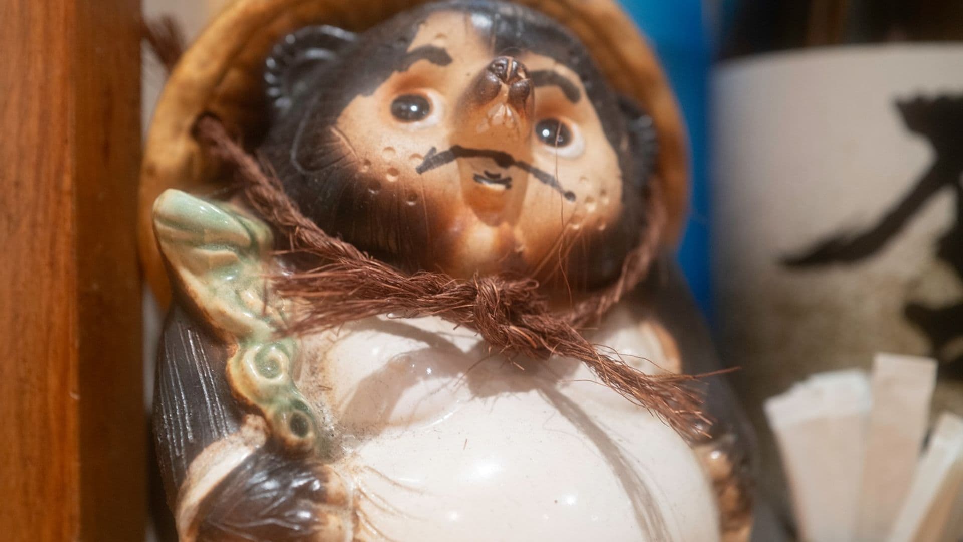 Decorative ceramic figurine of a smiling face, adorned with a hat and holding a leafy stalk, placed on a wooden surface among other decorative objects.