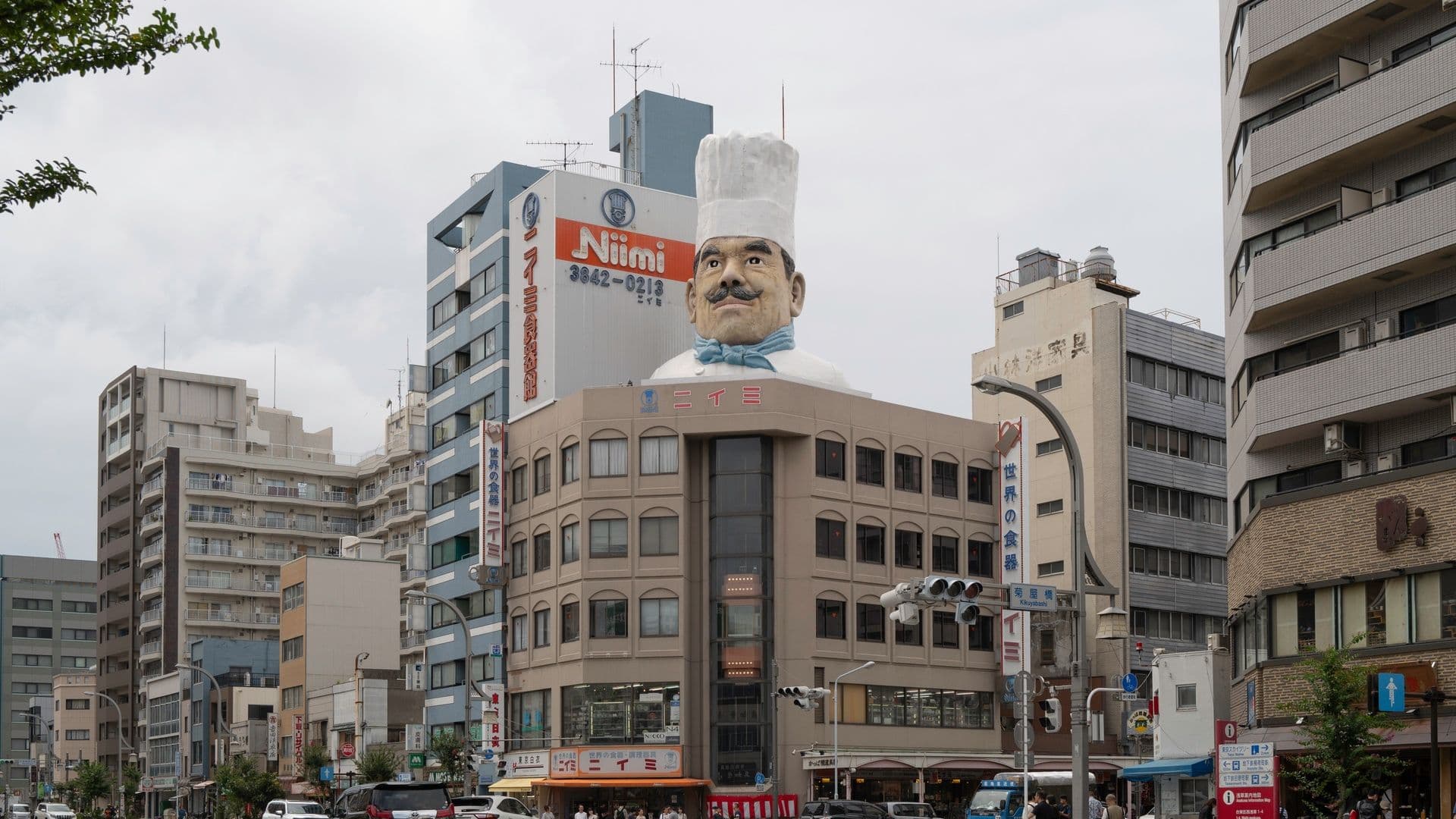 City street scene with a large building topped by a giant statue of a chef with a tall white hat, mustache, and blue neckerchief. The building has various signs with Japanese writing, and there are cars and pedestrians on the street.