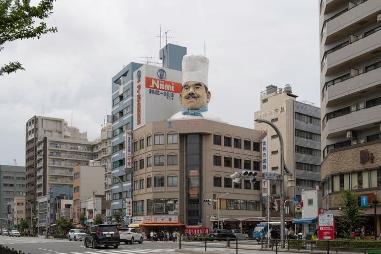 City street scene with a large building topped by a giant chef statue with a white hat and mustache, surrounded by modern multi-story buildings, cars on the street, and pedestrians.