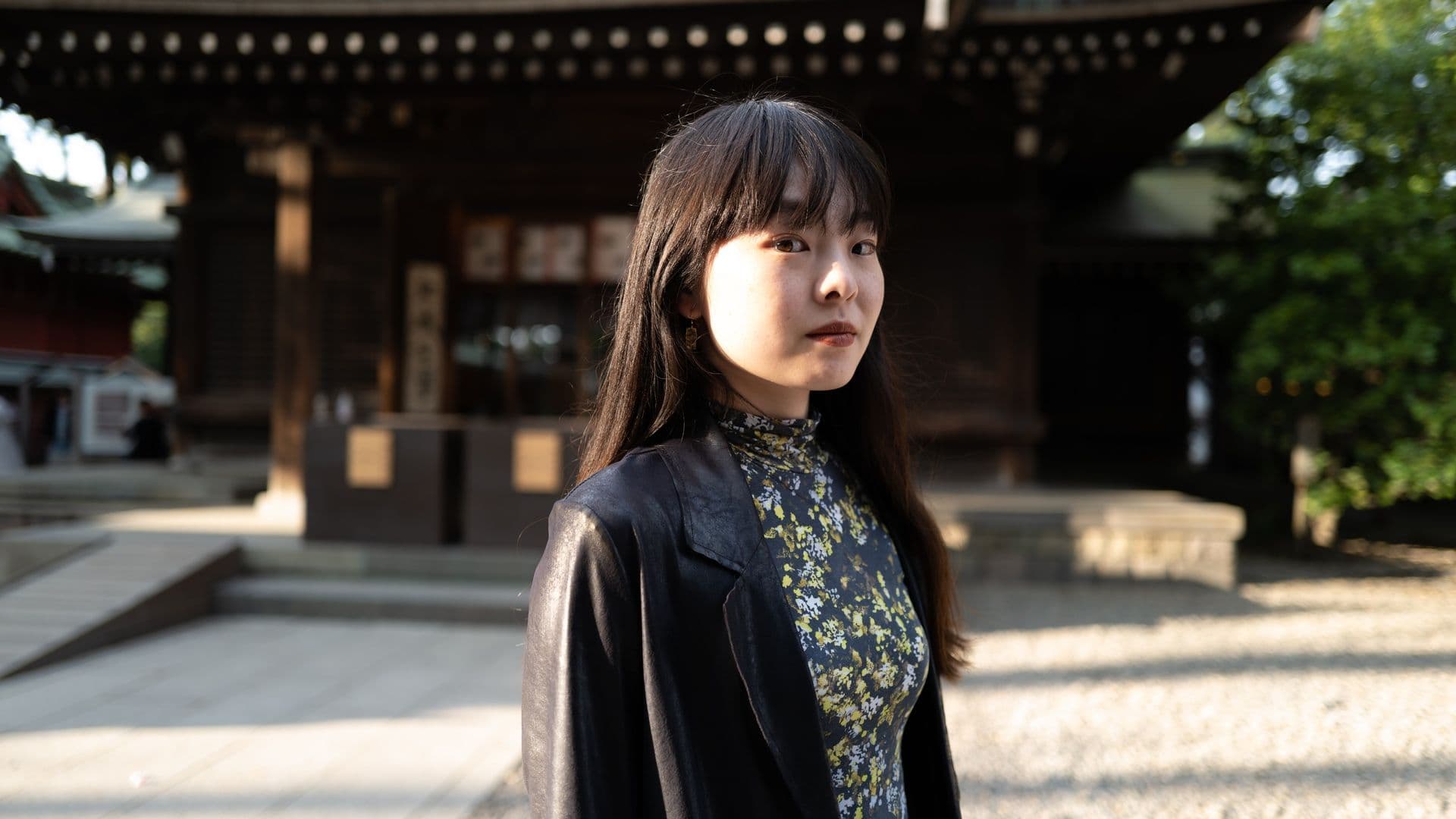 A young woman with long dark hair and a leather jacket stands outdoors in front of a traditional Japanese wooden building, illuminated by soft morning or late afternoon light.