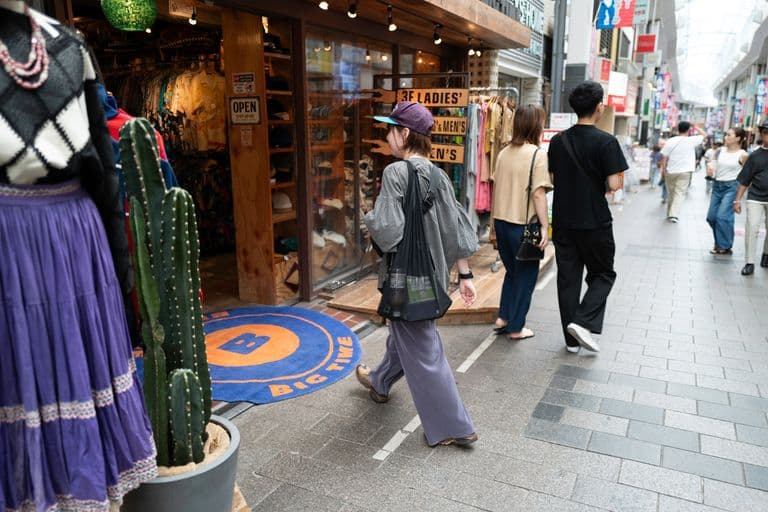 People shopping on a busy outdoor sidewalk with clothing stores and signs indicating sections for ladies, men's, and kids' clothing.