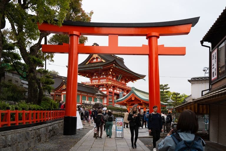 Fushimi Inari Shrine entrance with large red torii gate and tourists walking underneath in Kyoto, Japan.