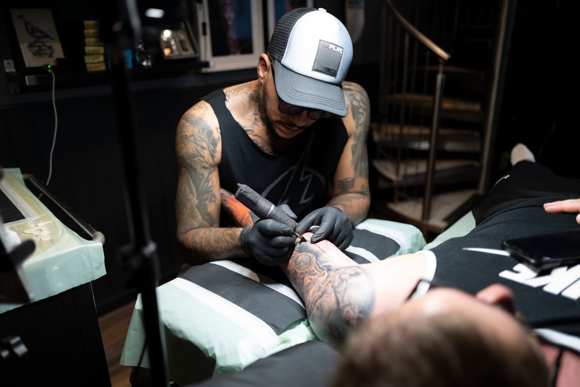 A tattoo artist is tattooing a person's arm in a tattoo studio, wearing a trucker hat and black gloves.