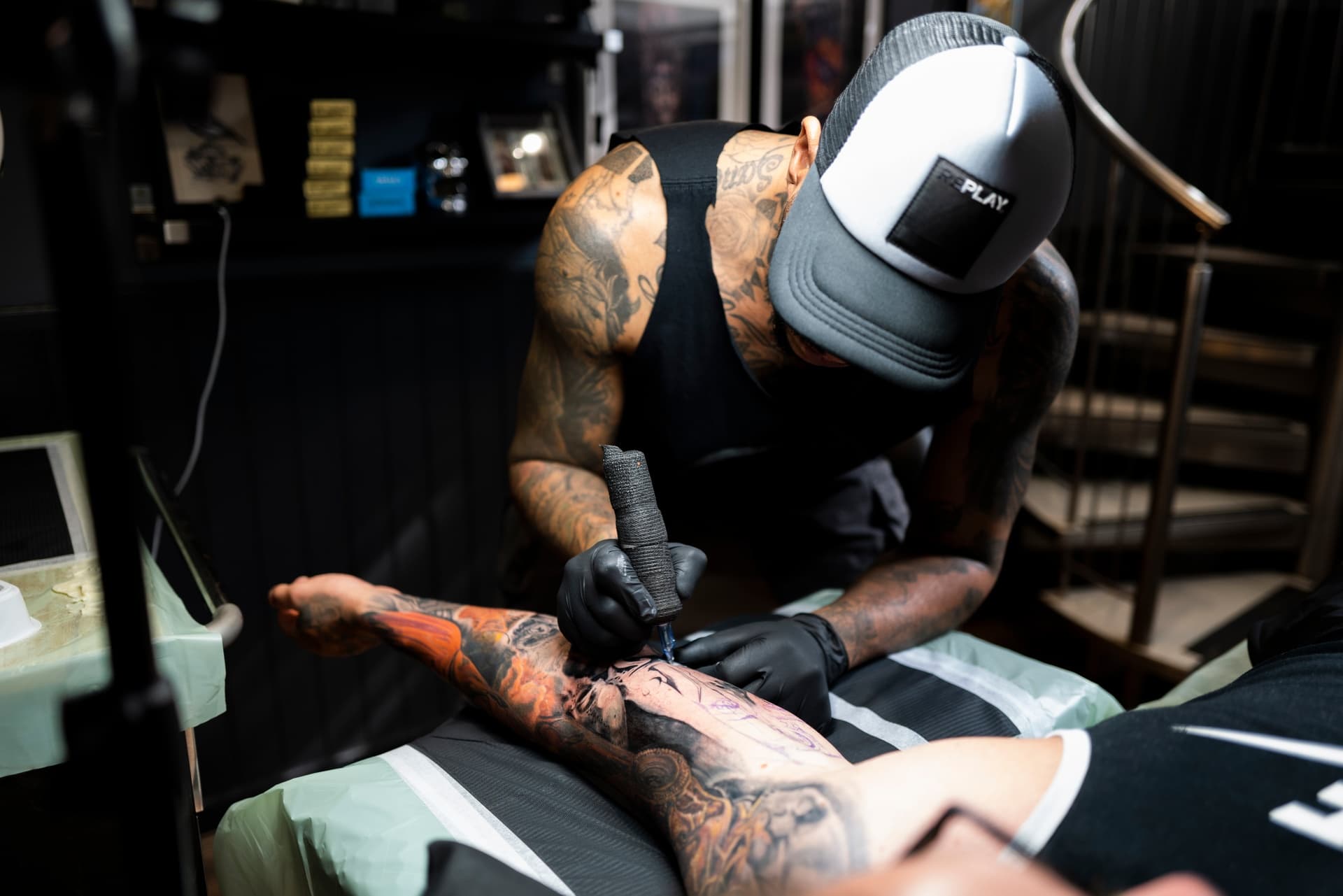 A tattoo artist is working on a person's arm in a tattoo parlor, using tattoo equipment. The artist is wearing a cap and gloves, and there are tattoo ink supplies in the background.