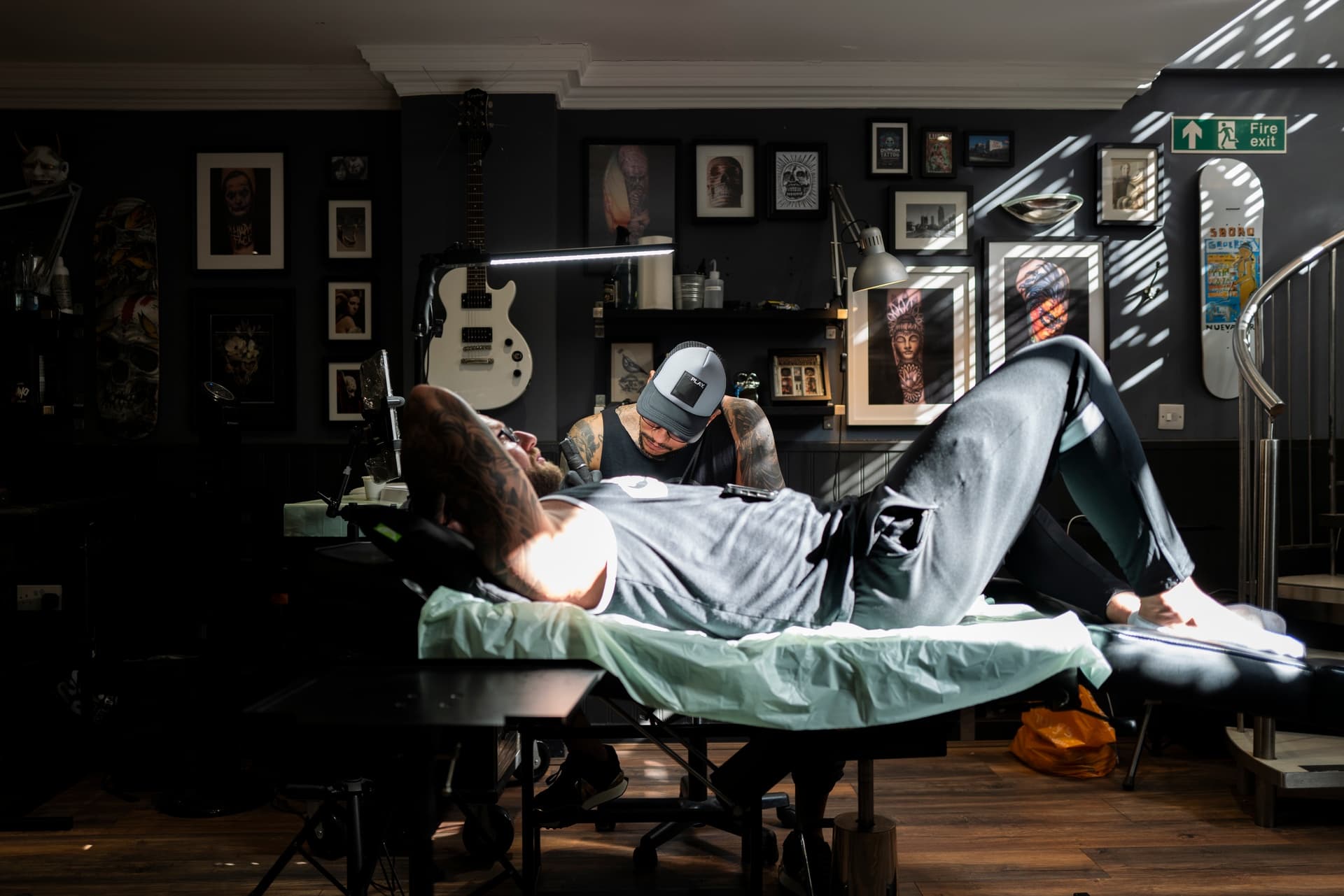 A man lying on a tattoo chair getting tattooed by an artist in a studio decorated with artwork, guitars, and tattoo-related items.