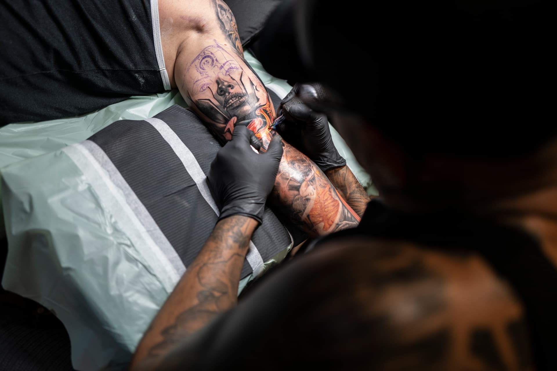 A person with tattoos getting a colorful tattoo on their arm from a tattoo artist wearing black gloves.