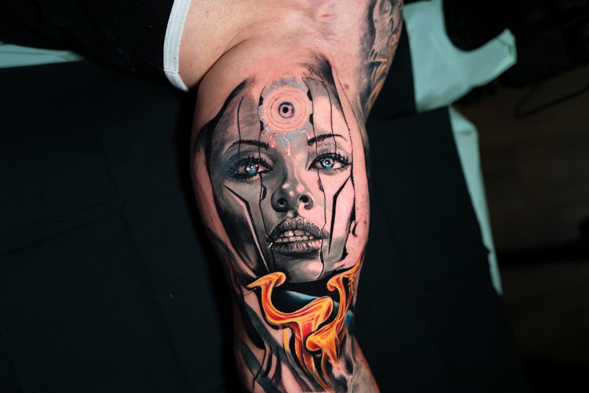 A person's thigh tattoo featuring a realistic portrait of a woman with blue eyes and black makeup, surrounded by abstract and fiery elements.